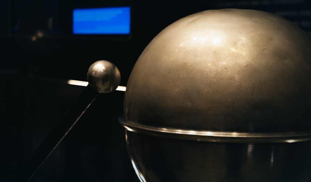 Esfera metálica polida em destaque, com esfera menor sobre trilho ao fundo em ambiente escuro de exposição.