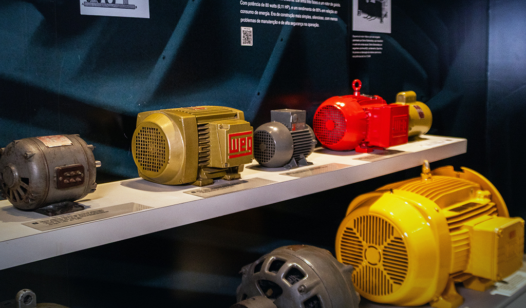 Motores elétricos antigos e modernos expostos em prateleira de museu, com etiquetas informativas.